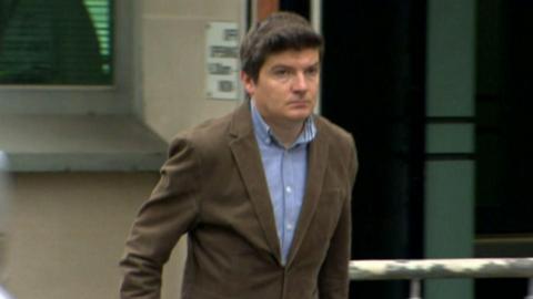 McMonagle walking outside a building. He has short brown hair, and he's wearing a brown suit with a blue shirt.