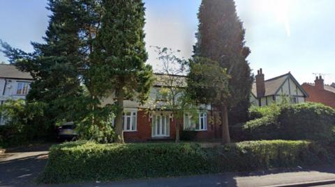 Buildings are in the background, with some trees and bushes nearer to the pavement in the foreground.