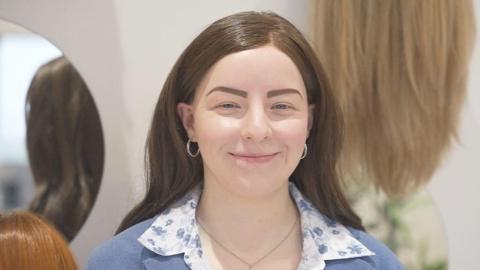 A picture of Megan McCready, who has alopecia, smiling. She is wearing a wig and is pictured in front of several other wigs