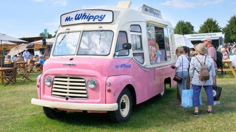 A vintage pink and white ice cream van is parked on a fild with a small queue snaking from the serving window.