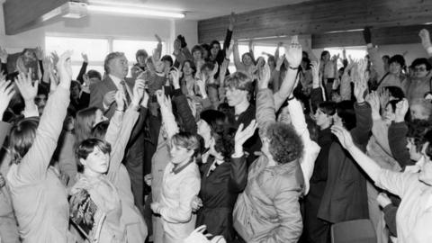 A black and white shot of factory workers packed tightly in a room, all with their hands in the air. Most are women wearing overalls.