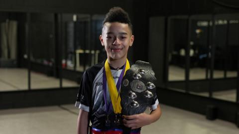 A boy in a mirrored studio smiles at the camera holding a trophy and wearing a belt that reads Oireachtas Rince na Gaeilge. He has tight curls cut into a mohawk style