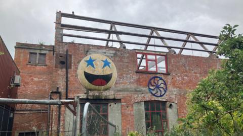 Part of the Life Factory building mid renovation with no roof and a battered smiley face on the side of the building. The sky is grey and there is greenery in the foreground.