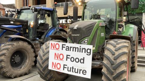 A green tractor parked in a square. There is a sign attached to its front that says "no farming, no food, no future"