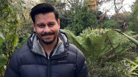 José Palma smiles at the camera, he is wearing a black puffer jacket and has a goatee and moustache. There are plants behind him.