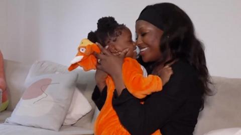 A young baby wearing an orange onesie is held up to her mother's face as the two exchange smiles