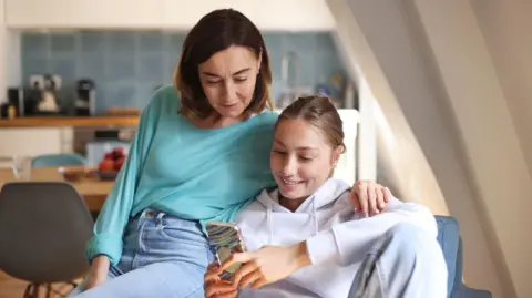 A mum and daughter sit together on a blue sofa in a bright room, looking at a smartphone. The mum, who has dark brown hair and wears a turquoise top with blue jeans, has her arm around her daughter's shoulder. The daughter has fair hair tied back and wears a grey hoodie and holds the phone. A kitchen with white cupboards, blue wall tiles, wooden worktops, chairs, appliances and a fruit bowl is blurred in the background.