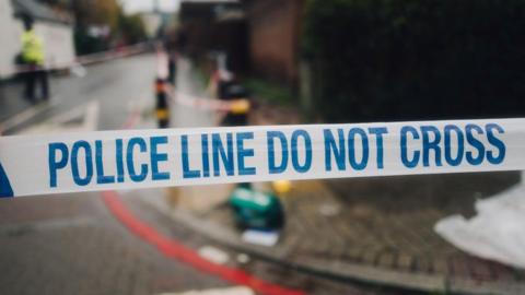 Police tape with a crime scene and police officers behind it on a road