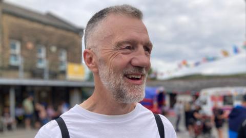A man with greying short hair and a beard stands laughing in the street.