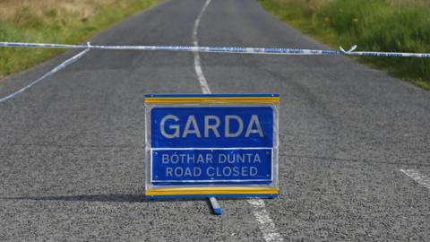 A stock image of a blue and white road sign on a rural road, warning drivers in both English and Irish that the road is closed. Above it there is a blue and white Garda (police) cordon stretching across the road.
