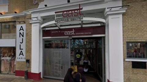 The exterior of the East Shopping Centre in Forest Green, London, with passers by outside. Signs are located above the doors which also show the name of the shopping centre.