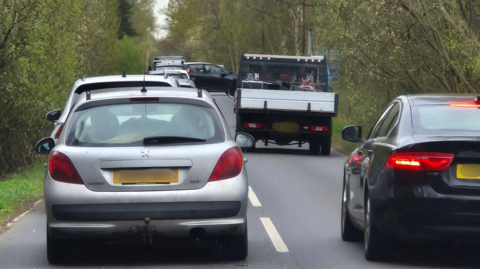 A line of traffic on a narrow, two‑lane road bordered by dense greenery. The road appears to be in a rural or semi‑rural area, with trees and bushes lining both sides and no visible buildings nearby On the left, there is a silver hatchback viewed from behind. The car has a yellow rear license plate. The brake lights are not illuminated.
On the right, there is a dark‑coloured (possibly black) sedan, also viewed from behind. The red rear lights on this vehicle are illuminated, indicating braking or slow movement. Both vehicles are positioned close together, suggesting slow or stopped traffic. Ahead of the foreground cars, there is a small flatbed or light lorry in the same lane as the silver hatchback. The lorry has an open rear bed with metal sides, and objects are visible in the back, though they are not clearly identifiable. Several other vehicles are visible further ahead, forming a queue that extends along the road.