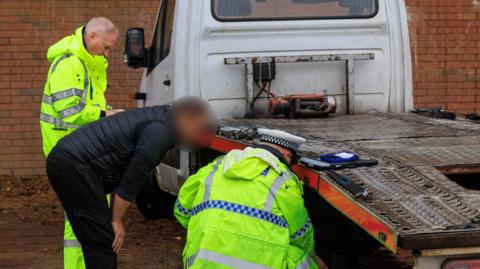Two police officers wearing yellow high-vis jackets and trousers look at the tyres of a flatbed truck. A man wearing a black puffer vest bends down next to one of the officers to look at the back tyre of the truck. His face has been blurred.