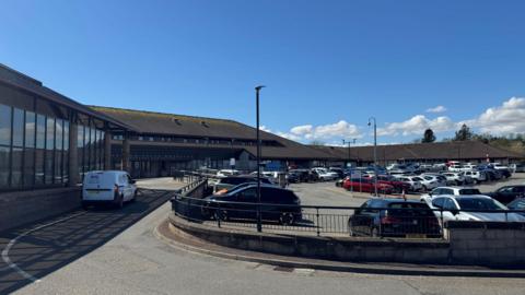 An exterior view of the front of Western Isles Hospital's main building. It is a low building with a peaked roof. There is a large car parking area outside the building with cars parked in it. A silver taxi is parked on a road in front of the main building.