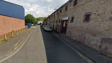 A Google street view image of Willway Street in Bedminster. The road is lined with industrial buildings, some of them with brickwork on the exterior. The picture is taken on a sunny day and there are some vehicles parked on the right-hand-side of the road