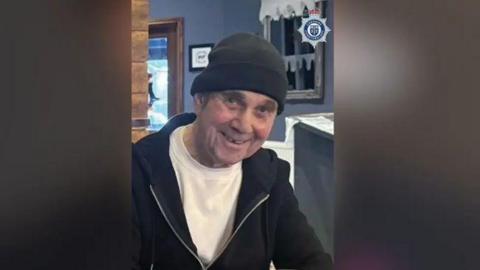 John McColl wearing a black hat and white t-shirt, with a black hoodie, is sitting indoors. 