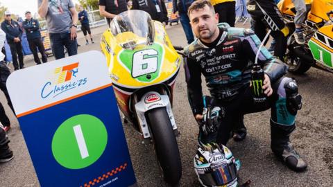 Sunlop kneeling next to the bike, which is yellow at the front with a white number six on a green plate. There is a Classic TT board with the number one on it next to the bike.