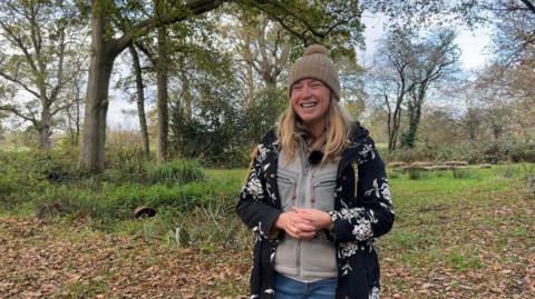A woman wearing a black and white cardigan and a beige woolly hat is standing in a forest and smiling
