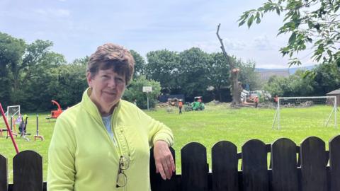 Val Beety, wearing a lime green jumper, leans on a fence. In the background, you can see a children's play park and some goalposts. Behind that, you can see the branchless elm tree while it is being felled.