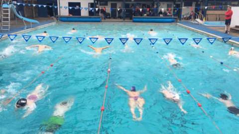 Swimmers in Stanhope Open Air Swimming Pool. Strings of bunting hang widthways over the pool and a lifeguard stands on the side of the pool. Lanes are marked out with ropes and buoys and the approximately 14 swimmers in shot all appear to be doing the butterfly stroke. At the far end of the pool a number of people sit or stand in front of a white building which has doors and windows with dark frames. On the left hand side of the pool stands a slide, much like a family with a child might have in their garden. A silver ladder leads to the top but the blue slide winds towards the pool.