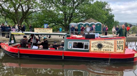 A canal boat with a group of musicians performing for the crowd in the background.