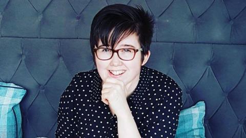 Lyra McKee stands with her right hand underneath her chin in front of a brightly painted blue wall. She is a young woman with short black hair, with a fringe over her forehead coming to rest on her glasses. She is smiling and wearing a black and white shirt.