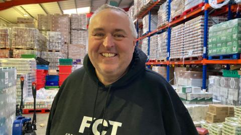 A man wearing a dark hoodie stands in a warehouse full of pallets containing food products.