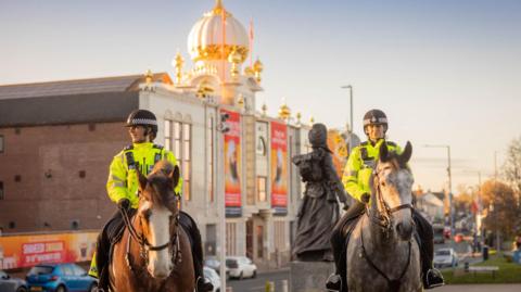 Two female police officers in uniform sit on horses which they ride past a gurdwara with a white and gold turret.