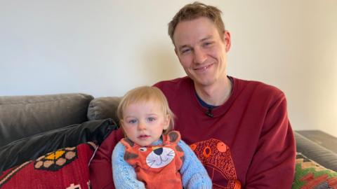 A man, wearing a red jumper, sits on a sofa with his son on his knee. The toddler is wearing a tiger outfit and has blonde hair.