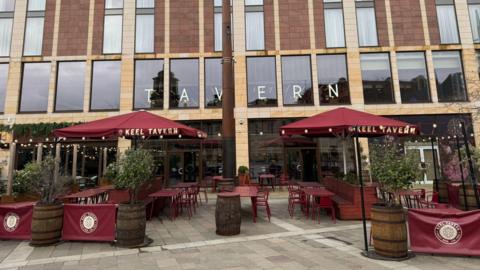 Outside of Keel Tavern bar. Large windows have lettering plastered on the insides which say "Tavern". Outside the building are red tables and parasols. The parasols say Keel Tavern