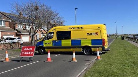 A yellow Cleveland Police Matrix van is parked across a road behind a road closed sign and traffic cones.
