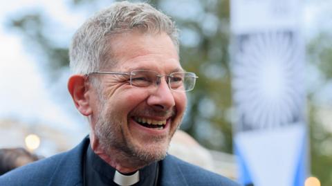 A man with short grey hair, glasses, a vicar's collar and a blue jacket, looks to his left and smiles.