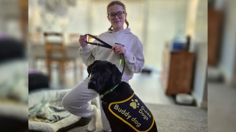 Aniyah holds onto Spirit's lead. She is a black lab wearing a Guide Dogs coat which says Buddy Dog. Aniyah is smiling