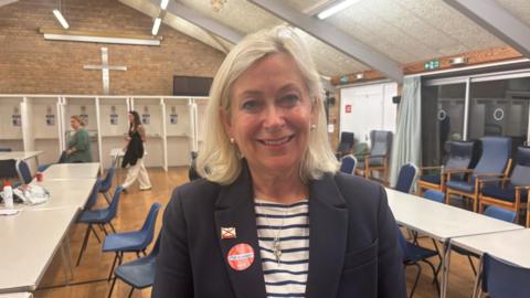 Helen Miles, a woman with shoulder length blonde hair and white earrings. She is wearing a white and navy stripy top underneath a navy blazer. On her lapel is a badge with a Jersey flag, and a sticker saying 'I'm a voter'. A number of tables are visible behind her in a church hall.
