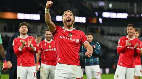 Wrexham striker Sam Smith punches the air in celebration with teammates applauding in the background
