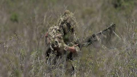 A British sniper wearing camouflage and a helmet covered in foliage takes part in training while in Kenya