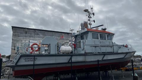 A grey ship, The Leopardess, behind a fence. It has the States of Guernsey logo on the side. 