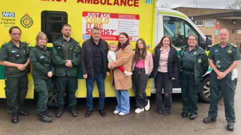 Ken Christy, his daughters and his partner Gemma Hurrell standing in front of a yellow ambulance and next to five members of ambulance staff in their dark green uniform.