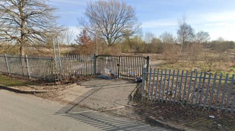 Derelict land, including a concrete roadway and grassland, is guarded by metal fencing and black double gates. A number of tall bare trees stand at intervals against a wintry blue sky. 