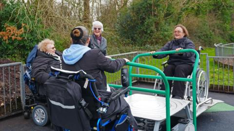 Three women in wheelchairs hold green bars attached to a piece of play equipment. A woman stands by them and smiles widely 