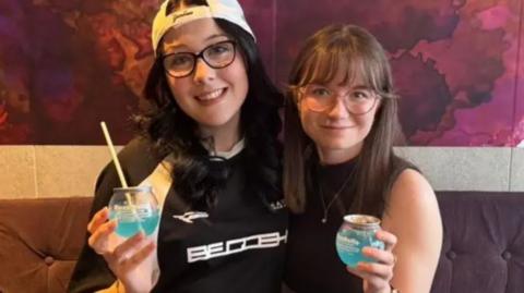 Caitlin and Katy sit on a purple sofa smiling while holding light blue BuzzBall canned cocktail drinks.