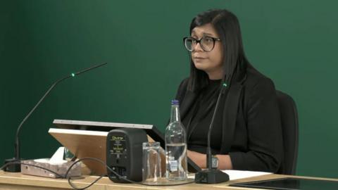 A woman sat at a desk in front of a screen and two microphones. She is wearing black clothing, has dark hair and black-rimmed glasses.