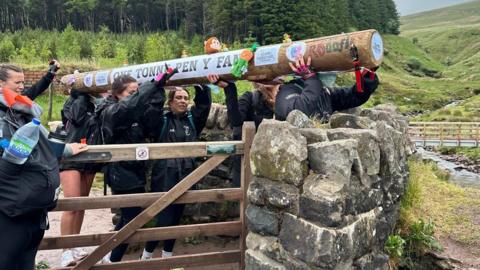 A group of women carrying a large wooden pole. They are walking through a wooden gate. The mountain is in the background, along with a wooded area.