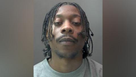 A police custody image of a defendant. He has slightly raised his head up and is looking directly at the camera. He is expressionless and has his eyes half closed. He has a dark, wispy, goatee beard and a moustache. His hair hangs in small plaits.