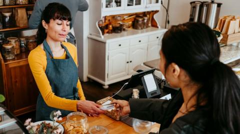 A person pays in a cafe with one barista taking payment, and the other fixing coffee behind the counter.