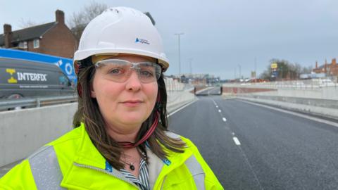 Shot of a woman wearing safety googles, hard hat and a yellow high-vis jacket. She is standing close to where the road leads down to an underpass.
