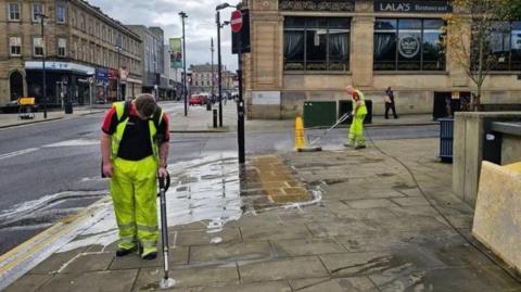 Two men in hi-vis clothing using power washers to clean streets of gum.