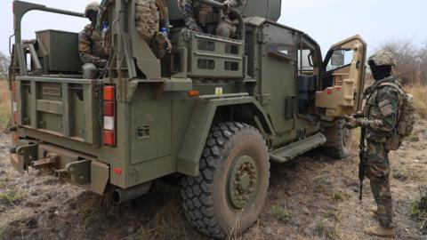 Ghanaian soldiers in camouflage uniform on a green military truck with the front door open and one soldier standing beside it 