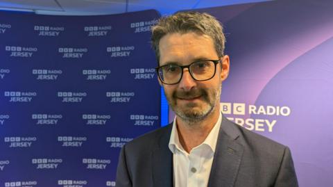 A man in a suit standing in front of a backdrop that repeatedly displays the text "BBC RADIO JERSEY."


