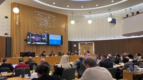 Round conference room with wooden panels on walls, people seated behind long tables with microphones, facing a wooden panel of people at front with screen behind them screening the meeting